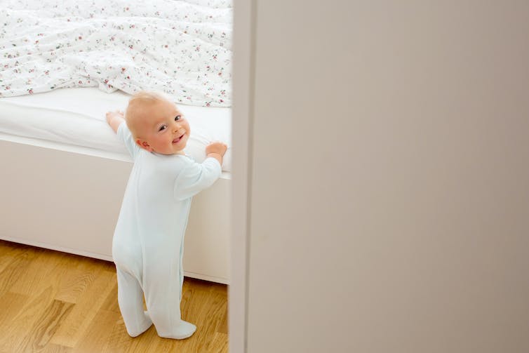 baby standing holding onto side of bed, looking back at camera