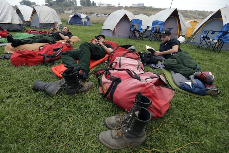Three firefighters lounge on air mattresses while reading. Tents are behind them, and boots are in the foreground.