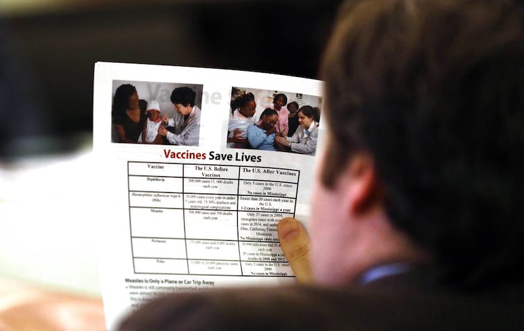 Over-the-shoulder shot of person holding a pamphlet reading 'Vaccines Saves Lives' with a table of the effects of different vaccines on the U.S. before and after their implementation