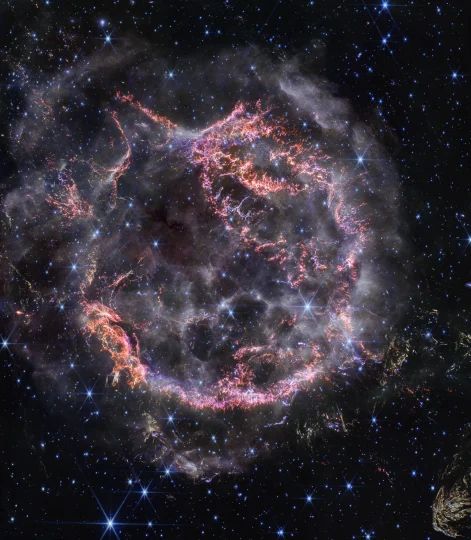 NASA’s James Webb Space Telescope’s new view of Cassiopeia A (Cas A) in near-infrared light is giving astronomers hints at the dynamical processes occurring within the supernova remnant. Tiny clumps represented in bright pink and orange make up the supernova’s inner shell, and are comprised of sulfur, oxygen, argon, and neon from the star itself. A large, striated blob at the bottom right corner of the image, nicknamed Baby Cas A, is one of the few light echoes visible NIRCam’s field of view. In this image, red, green, and blue were assigned to Webb’s NIRCam data at 4.4, 3.56, and 1.62 microns (F444W, F356W, and F162M, respectively).