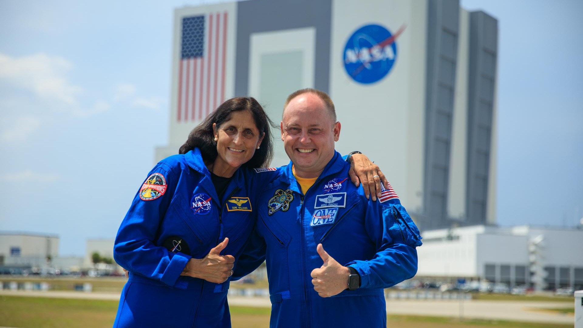 two astronauts in flight suits in front of a square shaped building far in back. the building shows the american flag and nasa logo on the side