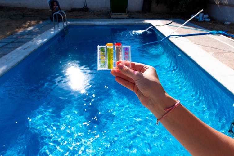 Hand holding pH and chlorine test kit above a swimming pool