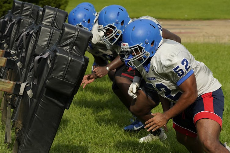 A line of linemen practice tackling while wearing helmets and pads.