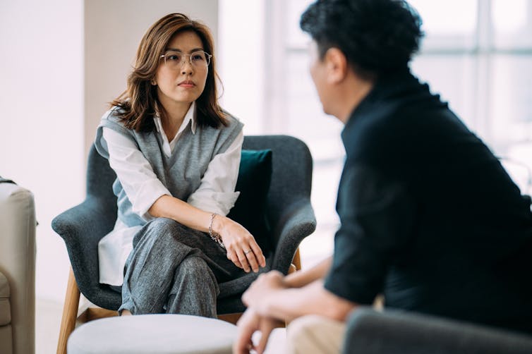 A woman with glasses, wearing a white shirt and sweater vest, sits listening intently to a man in a black shirt.