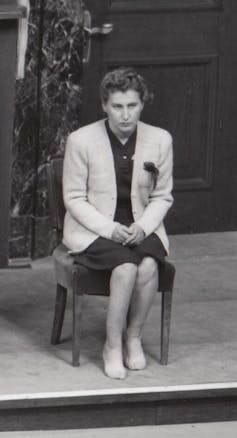 A woman with curly hair and a serious expression sits barefoot, wearing a skirt and suit jacket.