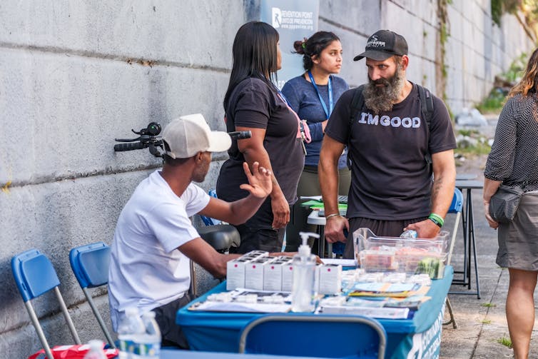 A man seated at table with harm reduction supplies speaks another man standing beside him