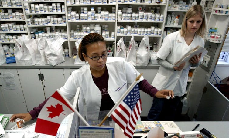 Two pharmacists behind the counter, shelves of drugs behind them and an American and Canadian flag before them