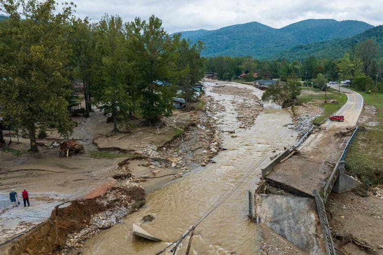 Two people stand near the edge of what once was a bridge across stream. The highway now ends at the edge and the bridge is broken and tipped into the water.