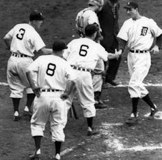 A baseball player at home plate, being congratulated by teammates.