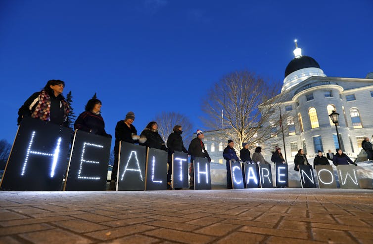 Line of people holding signs with individual letters spelling 'HEALTH CARE NOW,' standing in front of a white government building