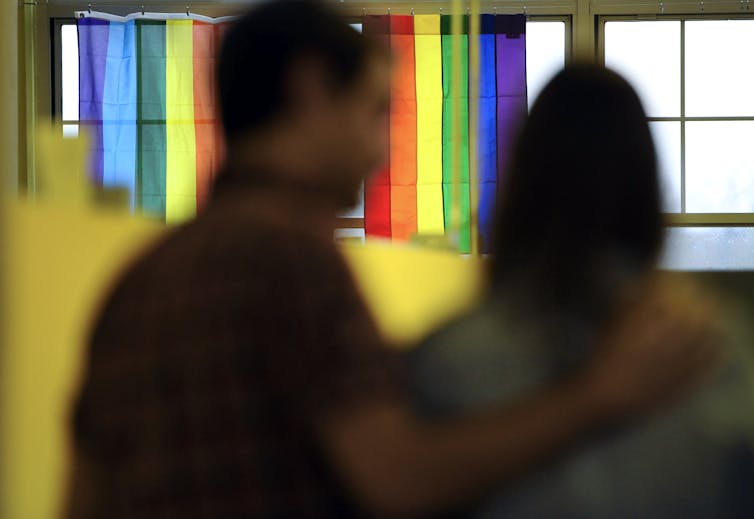 Person with hand on another person's shoulder, two rainbow flags hanging over a row of windows in the background