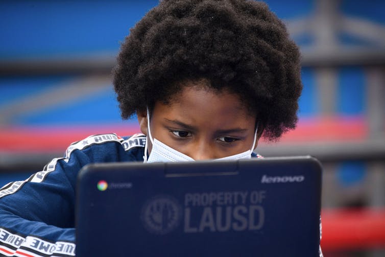 A child wearing a face mask looks at a laptop computer.