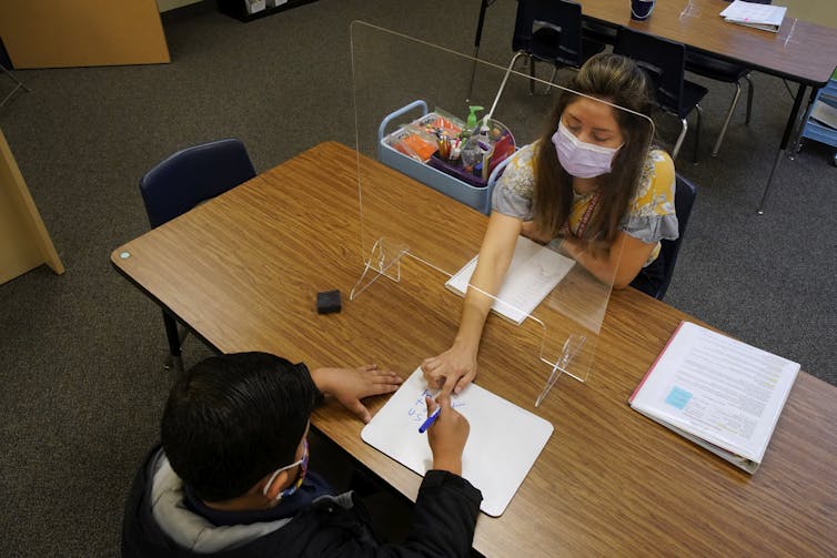 A teacher and student are separated by plexiglass as they sit across from each other at a desk.