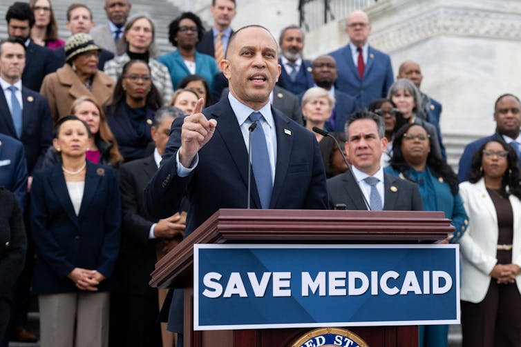 Man in a suit and tie speaks at an outdoor podium above the words 'Save Medicaid.'