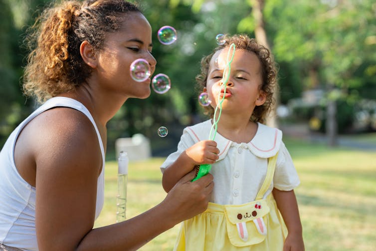 Young woman and a child blow bubbles together in a park.