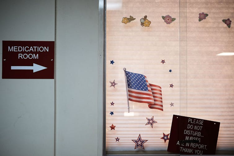 Sign reading 'MEDICATION ROOM' pointing to a window with stickers of an American flag, stars and flowers decorating it. At the bottom of the window is a sign reading 'PLEASE DO NOT DISTURB...NURSES ARE IN REPORT THANK YOU'