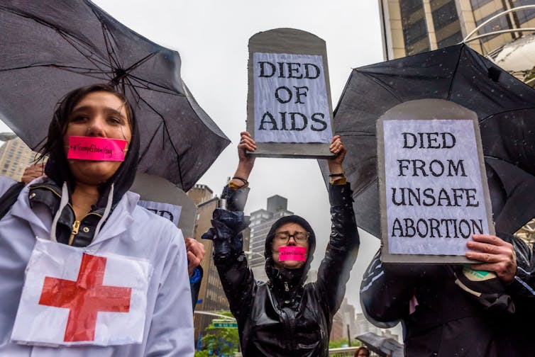 Protectors standing in the rain, holding grave-shaped signs reading 'DIED OF AIDS' and 'DIED FROM UNSAFE ABORTION,' with pink tape reading '#TrumpGlobalGag' over their mouths