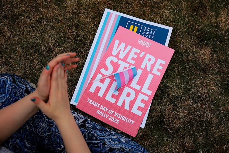 Hands clapping beside a small trans flag on top of a pile of signs, one reading 'WE'RE STILL HERE,'