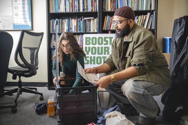 A man and woman pull papers from a collapsible cart while a poster behind them reads 'Boost Your Shot'