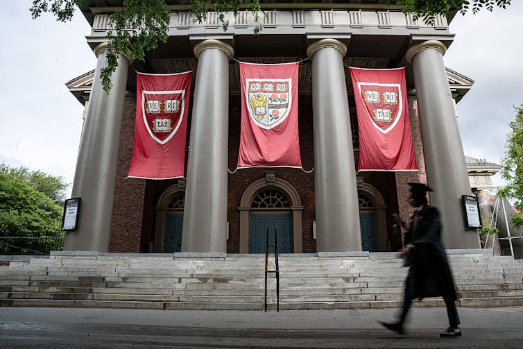 Building with Harvard insignia banners hanging between pillars, a student in a cap and gown walking past