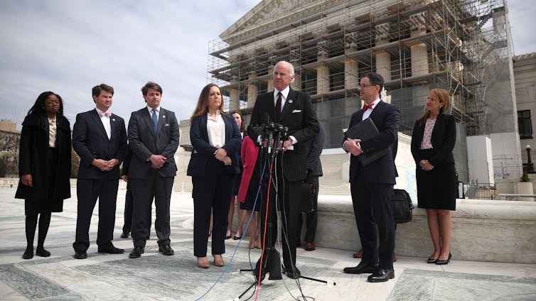 Man in suit speaks into a microphone, flanked by other people who are standing in front of a building surrounded by scaffolding.