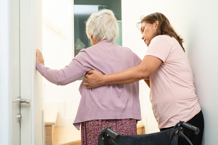 A caregiver helps support an elderly woman as she braces herself against two walls.