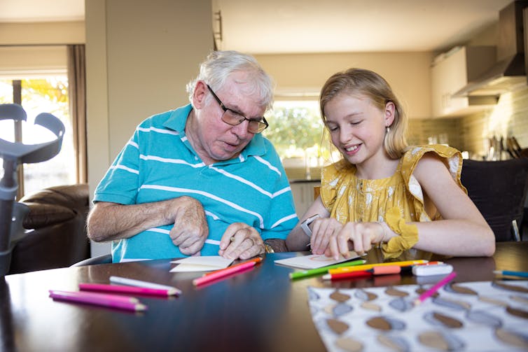 Senior man with dementia sitting at table with smiling young girl and colored pencils.
