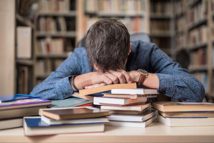 Person with head resting on forearms on top of a pile of books in a library