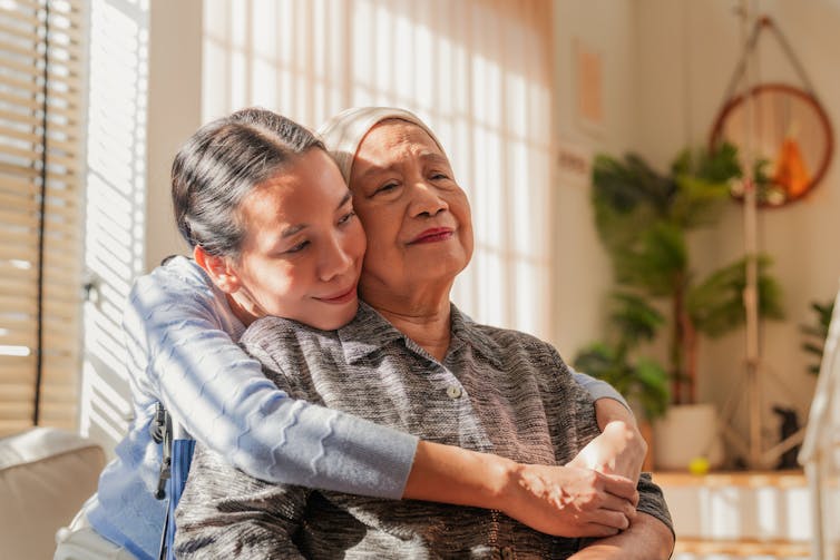 A young Asian woman reaches around to hug an older Asian woman from behind, as they sit in a sun-lit room.