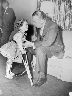 Young girl using crutches and leg braces smiles while walking to man in suit seated on a sofa