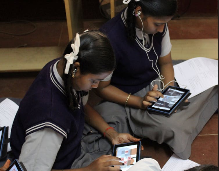 Two girls in a school uniform sit on the floor playing a game on a digital tablet.