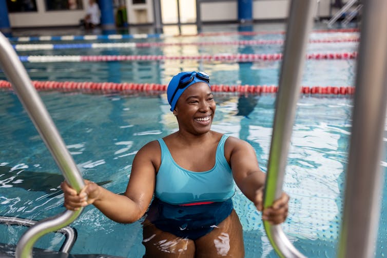 Smiling Black woman in swimsuit holding onto rails in indoor pool.