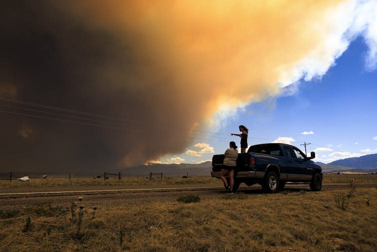 People stand in a field as a smoke plume turns the sky orange and then dark.