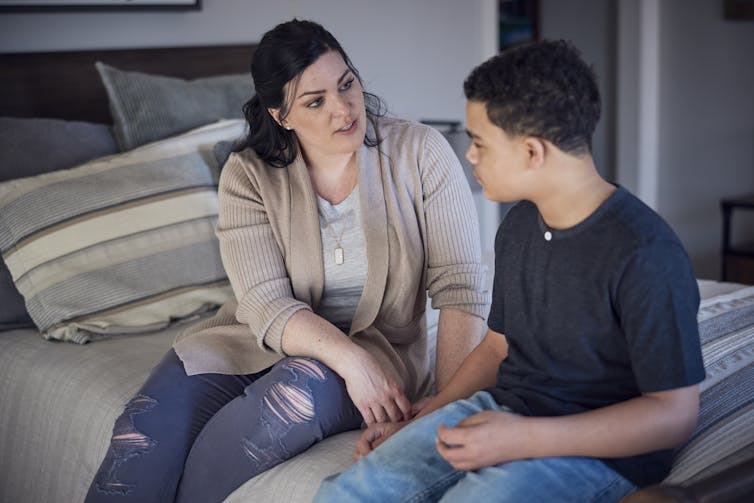 A middle-aged woman with a serious expression sits on a bed talking with a young boy.