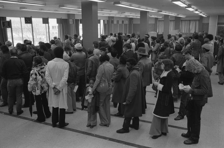 A large crowd of people wait on lines in a black and white photo from the 1970s.