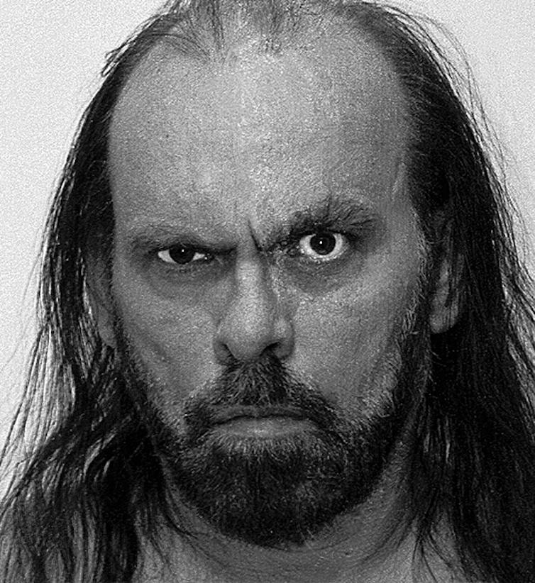 Black and white headshot of man with long hair and mustache and beard with one eyebrow raised